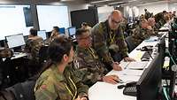 Soldaten verschiedener Länder sitzen in einem Saal in Reihen vor Computern. Im Hintergrund stehen digitale Bildschirme.