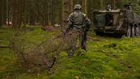 Ein Soldat zieht einen großen Tannenzweig im Wald hinter sich her, vor ihm steht geöffnetes Gefechtsfahrzeug.