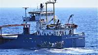 Ein graues Festrumpf-Schlauchboot fährt dicht neben dem Heck eines kleinen Frachtschiffs in See.