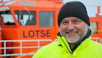 Ein Mann in wetterfester Kleidung mit verschränkten Armen; im Hintergrund signalrote Aufbauten eines Motorboots.