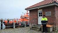 Ein Mann in wetterfester Kleidung vor einem kleinen Klinkergebäude; Im Hintergrund ein signalrotes Motorboot.