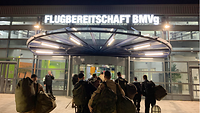 Mehrere Personen tragen Reisetaschen und befinden sich auf den Weg in ein Flughafenterminal