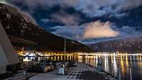 Panoramablick bei Nacht auf eine Küste mit beleuchteten Häusern, im Hintergrund verschneite Berge.