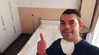 Ein Mann steht in einem Patientenzimmer eines Krankhauses. Er hält den ausgestreckten Daumen der rechten Hand hoch.