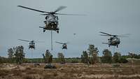 Vier Hubschrauber vom Typ NH-90 im Schwebeflug werfen Netze mit Material im Gelände ab.