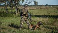 Ein bewaffneter Soldat geht mit einem Hund an der Leine im Gelände