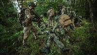 Mehrere bewaffnete Soldaten verschiedener Nationen stehen und knien um am Boden liegender Soldaten im Gelände.
