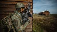 Ein Soldat steht mit seiner Waffe in der Hand an einer Hauswand. Im Hintergrund stehen mehrere Soldaten an einem Haus.
