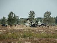 Mehrere Soldaten verlassen über die Heckrampe zwei Hubschrauber vom Typ Chinook und laufen zu Kettenfahrzeugen im Gelände.