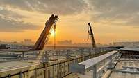 Mehrere Container und zwei Kräne stehen auf einem großen Gelände. Im Hintergrund geht die Sonne unter.