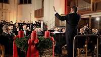 Männer und Frauen musizieren in einer Kirche. Im Vordergrund steht eine Adventskranz, auf dem eine Kerze brennt.