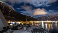 Blick von einem Schiff auf eine beleuchtete Hafenstadt vor Bergen bei Nacht. Am Himmel ziehen Wolken.
