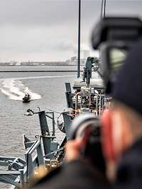 Ein Speedboot fährt auf ein graues Schiff zu; im Vordergrund verschwommene Umrisse eines Fotografen.