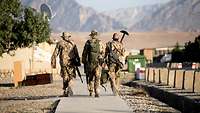 Drei Soldaten laufen nebeneinander auf einem Weg durch ein Bundeswehr-Camp. Im Hintergrund sind Berge zu sehen.