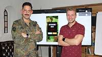 Ein Soldat in Uniform und ein Mann in rotem Hemd stehen vor einer digitalen Leinwand