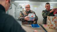 Ein Soldat greift nach einem sich auf einem Tresen befindlichen Paket. Neben und vor ihm steht jeweils ein weiterer Soldat.