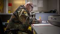 Frachtpapiere bearbeiten Ein Soldat sitzt an einem Tisch und füllt mit einem Kugelschreiber ein Blatt Papier aus.
