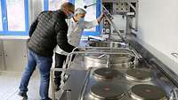Eine Frau und ein Mann in Arbeitskleidung begutachten einen Kochkessel in einer Containerküche