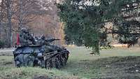 Ein gepanzertes Kettenfahrzeug steht auf einer Wiese. Zwei Soldaten sitzen auf dem Fahrzeug.