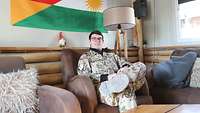 Ein Soldat sitzt auf einer Couch. Im Hintergrund hängt eine Flagge an der Wand.