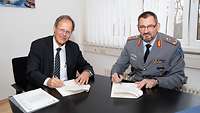 Zwei Männer sitzen an einen Tisch und Unterschreiben ein Papier