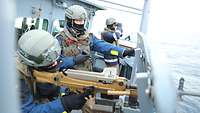 Drei Soldaten auf einem Schiff, einer schaut durch ein Fernglas, einer steht am Maschinengewehr, einer weist Richtung Meer