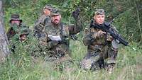 Fünf Soldaten knien mit Gewehren im Anschlag im Gras, einer eine Skizze in der Hand und zeigt in eine Richtung.