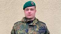 Ein Soldat mit grünem Barett steht vor einer hellen Häuserwand.