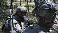 Zwei Soldaten liegen mit einem Funkgerät und Gewehren in einer Stellung im Wald.