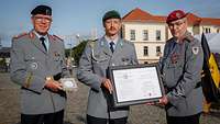 Drei Soldaten stehen auf einem Platz nebeneinander mit einer gerahmten Urkunde in den Händen.