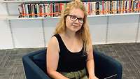 Eine junge Frau mit blonden, gelockten, langen Haaren sitzt auf einem blauen Sessel in einer Bibliothek.