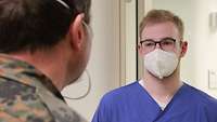 Ein Mann in blauem Kittel mit Mund-Nasenschutz steht einem Soldaten gegenüber.