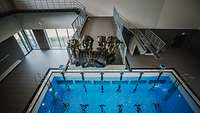 Vier KSK-Soldaten in einer Schwimmhalle mit einem zusammengerollten Schlauchboot auf einem Sprungturm über einem Schwimmbecken
