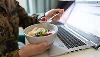 Ein Soldat sitzt vor einem Laptop und hält in der Hand eine Schüssel mit Obstmüsli und einen Löffel.