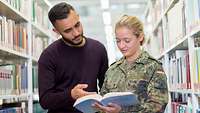 Ein Mann und eine Soldatin stehen in einer Bibliothek. Sie schauen gemeinsam in ein Buch.