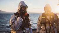 Zwei Soldaten mit ABC-Schutzkleidung und Atemschutzmasken stehen auf dem Deck eines Schiffes.