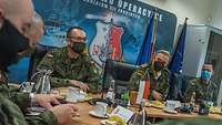 Mehrere Soldaten sitzen an einem Besprechungstisch; im Hintergrund ein militärisches Wandbild.