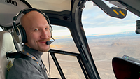 Ein Pilot sitzt in einem Cockpit. Er bedient die Steuerung. Der Hubschrauber ist in der Luft über einer felsigen Landschaft.