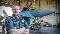 Ein Flugzeugwart in Fliegerkombination, mit vielen Aufnähern steht in einem Hangar vor einem Eurofighter