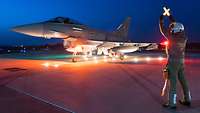 Ein Soldat steht neben einem Eurofighter bei Dunkelheit und hält ein leuchtendes Kreuz hoch
