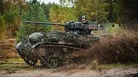 Waffenträger Wiesel 1 MK Ein Kettenfahrzeug Wiesel 1 MK fährt durch das Gelände und wirbelt Schlamm auf. Ein Soldat schaut aus der Luke des Fahrzeuges.