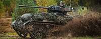 Ein Kettenfahrzeug Wiesel 1 MK fährt durch das Gelände und wirbelt Schlamm auf. Ein Soldat schaut aus der Luke des Fahrzeuges.