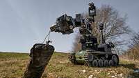 Ein ferngesteuerter Roboter hebt mit seinem Greifarm im Gelände einen Gegenstand vom Boden auf
