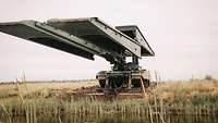 Ein Brückenlegepanzer Leguan steht mit ausgefahrender Brücke auf einer Wiese vor einem Gewässer.