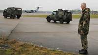 Ein Soldat steht auf einem Rollfeld, im Hintergrund zwei Militärfahrzeuge und ein Transportflugzeug.