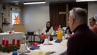 Ein Adventskaffee mit mehreren Menschen an einer eingedeckten Tafel, hinten an der Wand ein Kreuz