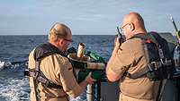 Zwei Marinesoldaten in sandfarbenen T-Shirts stehen an der Reling eines Schiffes; einer spricht in ein Handfunkgerät.