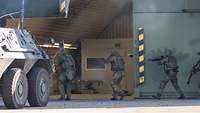 Vier Soldaten stürmen einen Flugzeugshelter unter Schutz eines Transportpanzers Fuchs.
