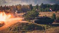Ein Kampfpanzer Leopard 2 steht hinter einem mit Gras bewachsenem Wall und feuert seine Kanone ab