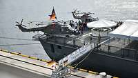 Eine Fregatte liegt am Pier, an Deck stehen zwei Hubschrauber und ein Getränkezelt